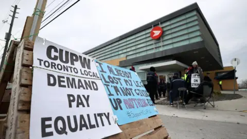 Canada Post workers picket outside Canada Post office as their strike continues in Toronto, Ontario on November 25, 2024. They put up posters outside their camp saying 'DEMAND PAY EQUALITY' and 'I ALREADY LOST MY LEG I'M NOT GIVING UP ANYTHING ELSE'.