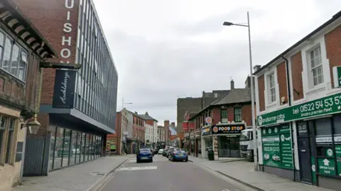 A view of Clasketgate in Lincoln. A large glass-fronted building is on the left, with a green taxi firm on the right. There is also a takeaway next to the taxi premises and cars are driving along the road.