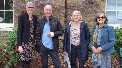 Ward Freman Community Group There are four people standing in a line in front of a building that has purple flowers on the walls and greenery. Left to right, the people are Joanne Willett, Philip Wylie, Alison Barr and Sheila White. They are all looking at the camera and smiling.