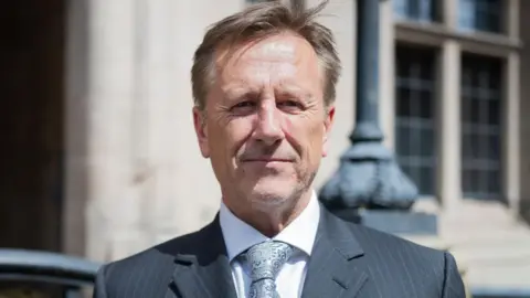 Martin Murray is standing infront of the ocunty council buildings in Stafford looking at the camera, he is wearing a grey tie, shite shirt and black suit jacket. 