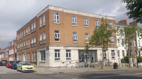 A three-storey building. There are cars parked to the left of the building. 