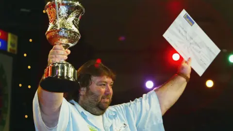 Getty Images Andy Fordham holding a trophy and a large cheque. He is wearing a white top