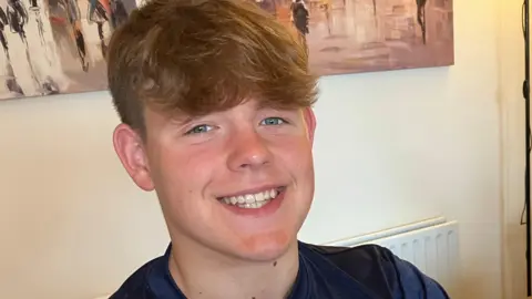 Oscar has brown hair pushed forward, he is smiling. He is wearing a navy blue T shirt with a light blue strip across the chest. He is sat by a table, on which is a certificate, three medals in blue cases and a large silver trophy. 