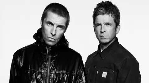 Black and white image of Liam and Noel Gallagher. Liam is on the left wearing a black shiny jacket and has short hair. Noel is on the right and wears a buttoned-up shirt.