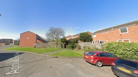 Residential street with red-brick houses, a small grassy area and tree, wooden fencing, and two parked cars, a red hatchback and a dark car, under clear blue sky.