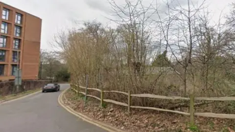 Hedgerow next to a road with a car on it and a red bricked building in the background.
