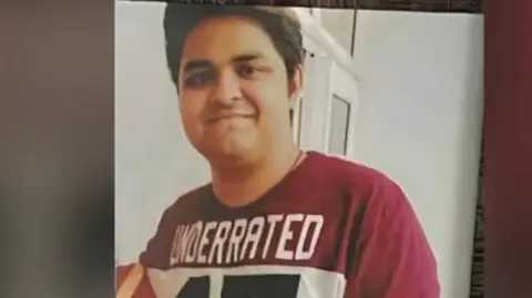 A photograph of Yuvraj Mehta - wearing a maroon-and-white tee shirt - posing for the camera, in a room with white walls