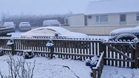 Five cars and vans parked on a residential street. Heavy snow has fallen and settled on their roofs and windscreens. There is a front garden fence in the foreground of the picture which is covered in snow. In the distance the sky is grey and foggy with low visibility. 