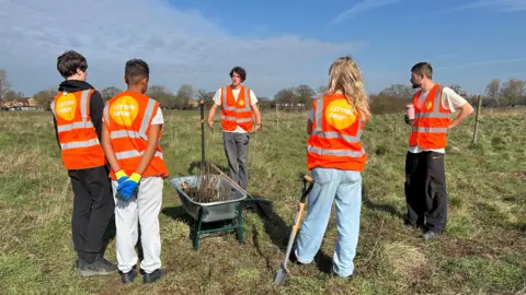 Letting Grow Children and young adults in orange hi-vis vests are gathered in a circle on a green patch of land around a metal wheelbarrow containing tree saplings