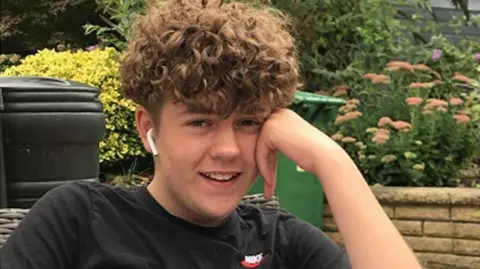 A smiling Olly Stephens with his head resting on his hand. He has curly brown hair and is wearing white earbuds and a black Nike T-shirt.