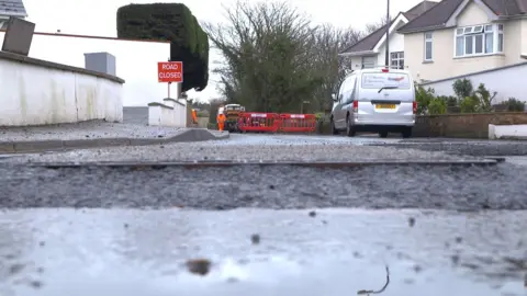 An eye-level of a road with a red sign in the distance. It has the words 'road closed' sign and two red barriers. There is the silver parked on the right as well as houses. There is a white wall to the left of the sign.