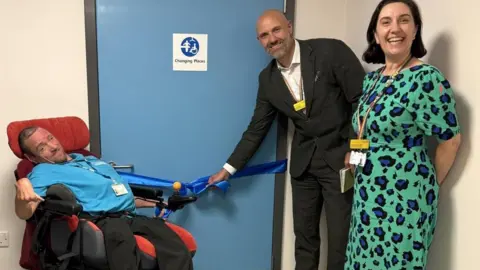 North West Anglia Foundation NHS Trust Matt, a man in a blue shirt, sitting in a red wheelchair. Chris Todd, a man in a black suit is standing next to him, holding a blue ribbon on the toilet door. A woman in a green and blue patterned dress stands nearby.