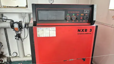 Vernon Lidstone A black and red oil-fired boiler in a utility room, surrounded by pipes and valves. The walls of the room are white.