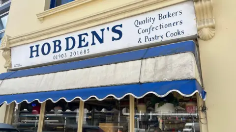 George Carden/BBC The Hobdens shop front which has a blue and white awning and signage which says "quality bakers confectioners and pastry cooks". 