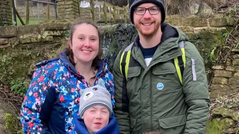 Kelsey Chappell Evan smiling at the camera with his mum and dad, Kelsey and Ash. They are standing outside in a garden and are wrapped up in warm winter clothing