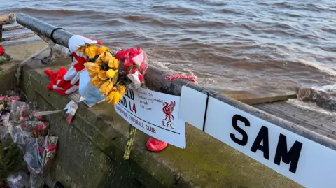Tributes pinned to a sea wall above the brown waters of a river estuary. They include red and yellow flowers, a red and white scarf, a Liverpool FC Anfield Road plaque and a car number plate that reads "SAM". 