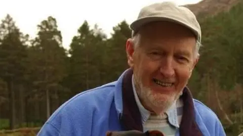 Hamish is wearing a cap and a blue fleece over a shirt and jumper. He is smiling.