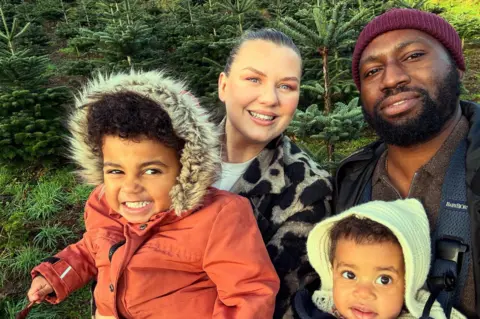 Patrick Penny-Annang A family of four taking a selfie - the kids are wearing warm winter clothes - and the man and woman are stood behind. Looks like they are in a Christmas tree farm.