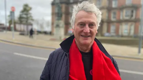 Mike Lloyd, a man with short white hair, wearing a red scarf and dark jacket and top, stands in a town centre and smiles.