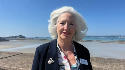 Debbie Taylor is wearing a dark blue blazer and a white shirt with floral patterns. She has white hair and is stood by the sea with Elizabeth castle in the background. 