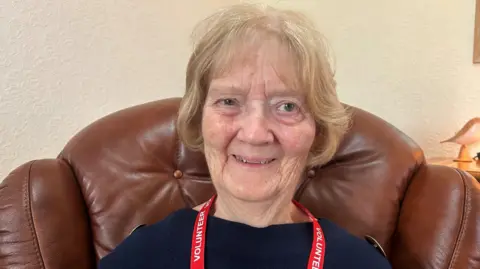 A close-up photo of 77-year-old Maureen Treharn. She has blond bobbed hair, and is wearing a red volunteer lanyard and blue jumper. She is sitting in a brown leather high back armchair and smiling at the camera. Only her head and shoulders are visible.