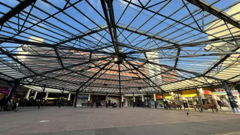 BBC A picture of the central precinct area of Anglia Square. It has an octagonal metal framework covering it, with various shops around the edges.