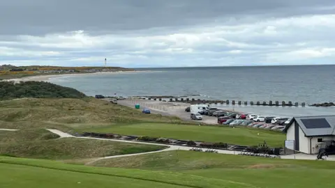 A wider view of a car park sandwiched between a golf course and the sea