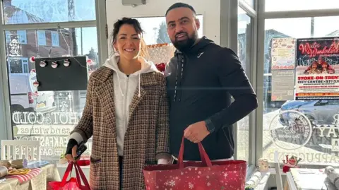 Samantha Taylor A man and woman are standing in a shop and smiling at the camera while holding two red Christmas-themed bags
