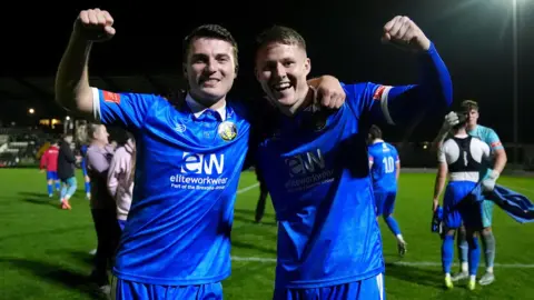 PA Media Two footballers celebrate with arms raised on a football pitch after a victory in the FA Cup. They are wearing blue shirts and shorts with a white sponsor's logo.