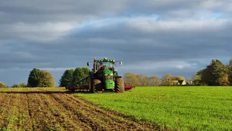 A tractor can be seen ploughing a field with a grey sky overhead. Half of the ground has been ploughed and the other half is covered in grass. A house can be seen in the distance.