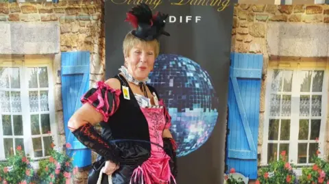 Family photo Pat is striking a pose in a flamboyant black and pink ballroom gown, fascinator and pink heels, in front of a popup stand branded with 'Strictly Dancing Cardiff'. 