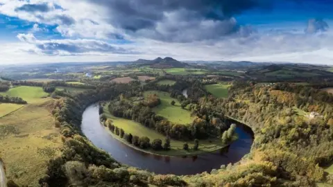 Jim Page The river Tweed on a big bend in the foreground with hills in the distance. The banks are heavily wooded but there are also many fields visible 