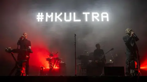 Getty Images Robert Del Naja and Grant Marshall on stage during the Massive Attack gig on The Downs in Bristol. The screen behind them is glowing red and black and the words "#MKULTRA" are visible in white letters