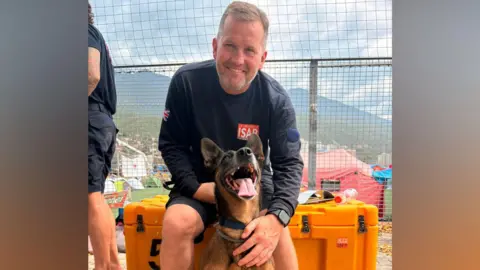 A man with light brown hair and a grey beard is smiling. He is wearing a long navy jumper with a red and white logo on it that reads "ISAR". He is sitting down and at his feet is a black and brown dog that has its mouth open with its tongue out.