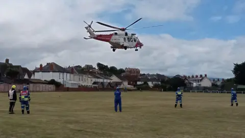 HM Coastguard A red and white helicopter landing on a playing field with rescuers standing on the ground