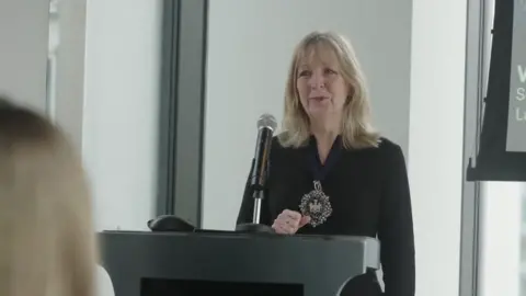 Dame Susan Langley, a blonde woman in a black shirt standing at a microphone giving a speech.