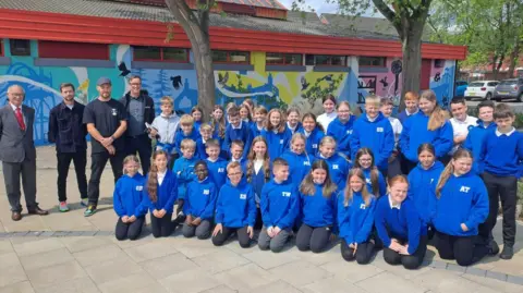 A primary school class all in blue jumpers. Three men stand on the left of the group. Behind them is a colourful mural featuring birds and tress.