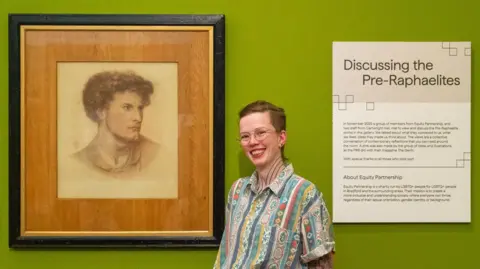A person wearing a colourful shirt with short hair stands next to a portrait of a man. On their right is a board that reads: Discussing the Pre-Raphaelites
