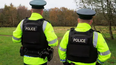 Two PSNI officers facing away from the camera, they are wearing black vests and high vis jackets.