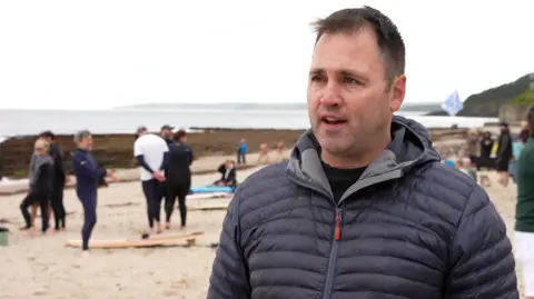A man in a navy blue coat. He has brown hair. Behind him are people stood on the beach. In the background is the sea, which appears grey, and the sky. 