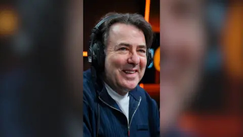 Broadcaster Jonathan Ross smiling with headphones on. He is wearing a blue jacket and white T-shirt.