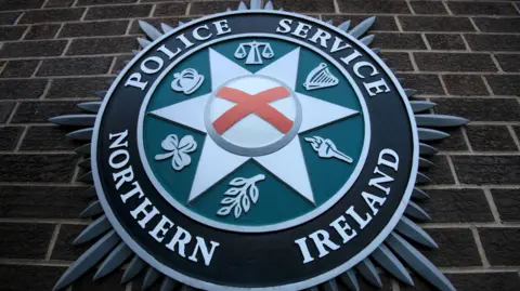 A Police Service of Northern Ireland crest on a brown brick wall.  The star-shaped crest consists of a red cross of St Patrick inside a central six-pointed silver star, which is inside a green circle, inside a larger black circle which bears the name of the service.  There are six emblems in the green circle, including a harp, a torch, an olive branch, a shamrock, a crown and the scales of justice.