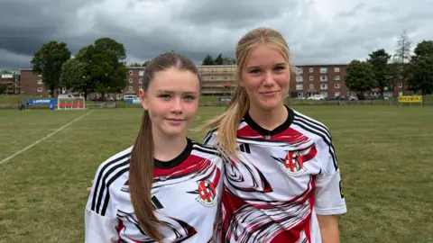 Two girls in Crusaders FC away shirts (white with red and black markings) stand on a football pitch. Low-rise flats and trees are in the background. Rachael has a long brown ponytail and is shorter than Daisy who has a long blonde ponytail.