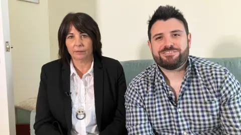 Elaine Pickford and her son Liam are sitting on a sofa. Elaine is wearing a black suit jacket and Luke is wearing a checked shirt.