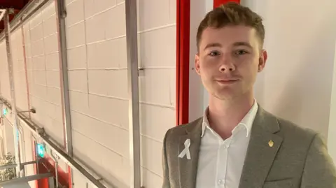 Amy Holmes/BBC A picture of a young man with short brown hair who is wearing a grey jacket with a white ribbon on it.