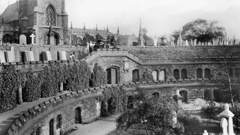 Heritage England A black and white photograph showing a funeral taking place at Warstone Lane. The church sits at the top of two tiers of catacombs and a small group of people are seen crowded at the centre of the catacombs in front of the church. 