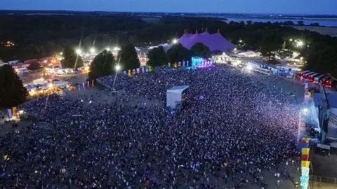 Martin Barber/BBC A aerial image of the Latitude festival's main arena in the evening, showing the audience, stalls and a side-one view of the stage. Woodland surrounds the site and the River Blyth estuary is off the festival site.