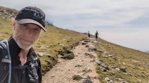 Andrew Lamb Andrew Lamb in selfie walking up mountain in sunny conditions. He is on the left of the image, wearing a black t-shirt and black cap. There are two people walking in front of him. He is in focus, while the two other people are blurred. 