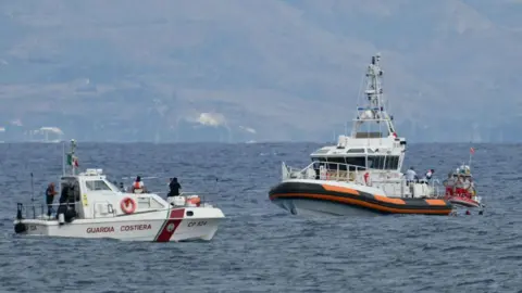Getty Images Three white and red Italian coastguard boats in a large stretch of ocean with land in the background. Three divers are in the water next to one of the boats.