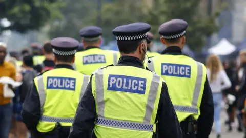 EPA Four uniformed police officers, wearing yellow hi-vis vests and black hats with a black and white checked band, patrol at Notting Hill Carnival on 27 August 2023